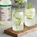 A bottle of Hometown Provisions Mojito Mix and two glasses of mojito garnished with lime and mint on a wooden board.