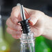 A hand placing a metal and rubber pour spout into the neck of a clear glass bottle.