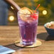 An Acopa Memphis highball glass of purple liquid with a cherry and orange slice on a table with a bowl of popcorn and a coaster.