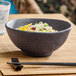 A dark gray round melamine bowl filled with food, placed on a bamboo mat with chopsticks and a glass of water nearby.