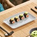 A light gray rectangular melamine platter displaying four pieces of sushi, shown on a bamboo placemat.