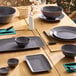 A set of dark gray round melamine plates arranged on a wooden dining table with matching bowls and tableware.
