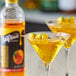 A glass of yellow liquid with a straw and fruit on top next to a bottle of DaVinci Gourmet Mango Fruit Syrup.