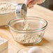 A glass bowl being filled with whole grain oat flour from a metal measuring cup.