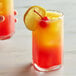 A hand holding a glass of red liquid with an orange drink on the table.