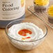 A large container of Hometown Provisions egg yellow food coloring is shown next to a bowl of white frosting with yellow coloring added, and cupcakes with yellow frosting in the background.