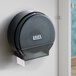 A black plastic jumbo toilet tissue dispenser mounted on a restroom stall wall.