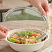A clear compostable plastic lid being placed on a wide fiber take-out bowl filled with salad.