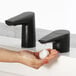 A person using a Bobrick matte black deck-mount automatic foam soap dispenser at a white sink with a black faucet.