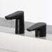 A Bobrick matte black deck-mount liquid soap dispenser next to a black faucet on a white counter.