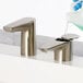 A hand in a blue glove using a Bobrick brushed nickel deck-mount automatic foam soap dispenser to pour soap into a sink.