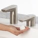 A close-up of a Bobrick brushed nickel deck-mount automatic foam soap dispenser being used to wash a person's hands.