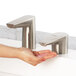 A person using a Bobrick brushed nickel deck-mount automatic soap dispenser to wash their hands.