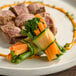 A bundle of assorted vegetables including carrots, green beans, and broccoli wrapped in a thin slice of zucchini, served on a plate with sliced meat in the background.