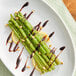 A plate of cooked asparagus spears garnished with pine nuts, lemon slices, and a drizzle of balsamic glaze.