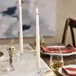 A pair of tall, white taper candles burning in clear candle holders on a set dining table.