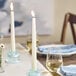 A white 10-inch taper candle burning in a glass holder on a set dining table.