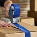 A roll of blue acrylic carton sealing tape being applied to a cardboard box with a tape dispenser.
