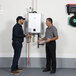 Two men standing next to a Rinnai tankless water heater with a white rectangular recirculation pump.