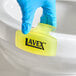 A yellow Lavex citrus scent toilet bowl clip being placed on the rim of a toilet by a gloved hand.