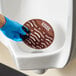 A brown Lavex EVA deodorizing gel urinal screen being placed in a white urinal by a person wearing a blue glove.