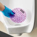 A round, purple Lavex deodorizing gel urinal screen being placed in a white urinal by a person wearing a blue glove.