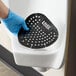 A black vinyl deodorizing urinal screen with holes, being placed in a urinal by a gloved hand.