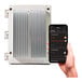 A hand holding a smart phone next to a white Bromic Heating Smart-Heat dimmer controller.