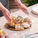 A clear round plastic high dome lid being placed on a catering or deli tray filled with assorted pastries and fruit.