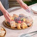 A clear round high dome plastic catering tray lid being placed over a tray of assorted pastries and fruit.