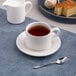A white embossed porcelain tea cup with a saucer, filled with tea and placed on a blue placemat.