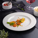 A white round porcelain plate with an embossed wide rim, featuring a small serving of vegetables and garnishes in the center.