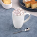 A white embossed porcelain mug filled with a whipped cream-topped beverage, placed on a blue placemat with a spoon nearby.