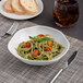 An Ariane deep porcelain plate with a bowl of pasta with tomatoes and basil on a table.