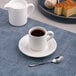 A white embossed porcelain espresso cup with a saucer and spoon, accompanied by a milk jug and desserts in the background.