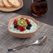 A bowl of Ariane Earth Dark Sand Dots porcelain filled with yogurt, fruit, and a spoon on a table with a glass of brown liquid.