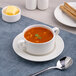 A white porcelain soup bowl with handles, filled with tomato soup and placed on a matching saucer.