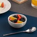 A small white embossed porcelain bowl filled with fruit, placed next to a spoon.