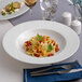 A round, wide-rim deep white porcelain plate with a pasta dish served in the center.