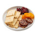 A white round melamine plate containing crackers, dried cranberries, dried apricots, and slices of cured sausage.