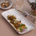 A white rectangular porcelain plate with food on a table with a fork and knife.