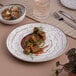 A white Ariane Tornado porcelain plate with food on a table with a fork and a glass of wine.