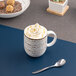 A white Ariane porcelain mug of coffee with whipped cream and caramel sauce with a spoon.