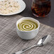 An Ariane Linen Dots porcelain bowl of soup with a swirl of cream in it on a table with a spoon and a glass of brown liquid with ice cubes.