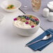 A large warm white porcelain bowl filled with yogurt, granola, blueberries, and edible flowers.