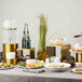 A three-piece set of square gold stainless steel risers displayed on a buffet table with various food items and decorations.