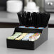 A black plastic coffee condiment organizer with nine compartments holding stirrers, creamers, sugar packets, and other coffee accessories.