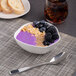 A white porcelain bowl filled with purple smoothie, blueberries, and nuts on a table with a spoon and a glass of brown liquid.