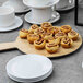 A round faux wood melamine serving board with a handle, displayed with pastries and surrounded by white cups and plates.