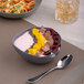 A bowl of fruit and yogurt with a spoon on a table with a glass of liquid.